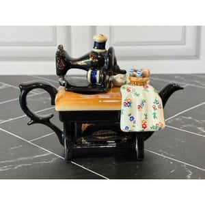 May include: A ceramic teapot shaped like a vintage sewing machine. The base is black with gold accents and the word INFUSIONS. It features a tan tabletop, a black sewing machine top, and a white cloth draped over the side with a colorful floral pattern.