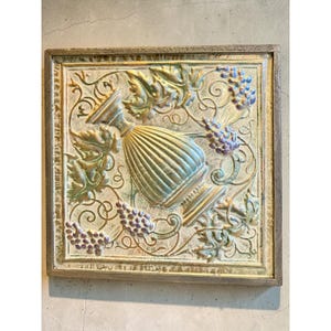 May include: A square wall plaque featuring a central embossed urn surrounded by grape clusters and vine leaves. The piece has a metallic finish with muted gold, green, and purple tones, all encased within a simple, weathered wooden frame.