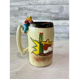 May include: A ceramic mug with a cream-colored speckled finish featuring a hand-painted green cactus and a figure wearing a yellow sombrero. A three-dimensional figure wearing a colorful hat is attached to the top of the handle.