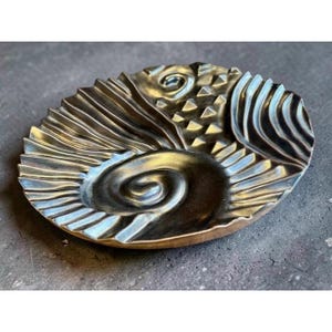 May include: A circular metallic dish with a dark bronze finish, featuring a textured surface with a prominent central spiral, radiating linear grooves, and a section of small raised triangular shapes across the top portion of the object.