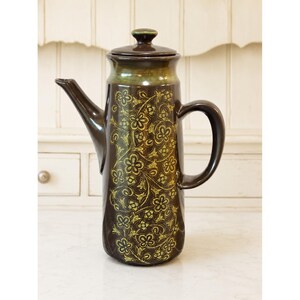 May include: A tall, cylindrical dark brown ceramic coffee pot with a matching lid. The body features a repeating yellow-green floral pattern. It has a curved handle on the right side and a pointed spout on the left, with a subtle green glaze near the top.