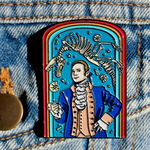 Puede incluir: Un pin de esmalte colorido con el retrato de un hombre con una chaqueta azul y una corbata blanca, sobre un fondo azul con un esqueleto de dinosaurio y otros fósiles. El pin está enmarcado con un borde de arcoíris.