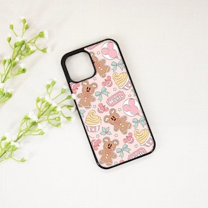 May include: A pink phone case with a black border featuring a repeating pattern of gingerbread men, cupcakes, bows, and Mickey Mouse silhouettes. The word "Magic" is printed on a label. The case is on a white surface with white flowers.