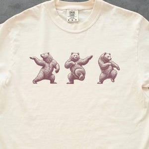 May include: Ivory-coloured t-shirt with a crew neck and short sleeves. The shirt features a graphic of three bears in various dance poses in a dark purple colour. The words "comfort colours" are printed at the top.