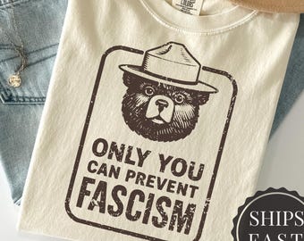 May include: Ivory-colored t-shirt with a graphic of a bear wearing a ranger hat and the text "ONLY YOU CAN PREVENT FASCISM." The shirt is laid on top of light blue jeans, a tan hat, and a necklace.
