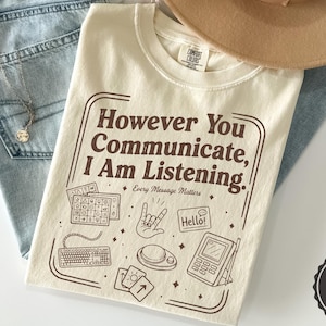 May include: An ivory t-shirt featuring the brown text 'However You Communicate, I Am Listening' and 'Every Message Matters' with illustrations of a communication board, a keyboard, a hand sign, a speech bubble, a switch, and a tablet device.