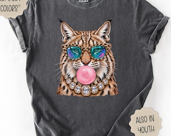 May include: A charcoal gray short-sleeve t-shirt featuring a graphic of a lynx wearing leopard-print glasses and a jeweled necklace while blowing a pink bubblegum bubble. The shirt includes a Comfort Colors label and text stating Also in Youth.