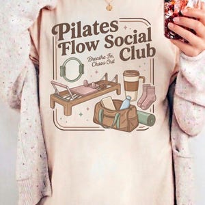 May include: Cream-coloured t-shirt featuring a Pilates Flow Social Club design. The graphic includes a Pilates reformer, a gym bag, a coffee cup, socks, and a yoga mat. The text "Breathe In, Chaos Out" is also included.