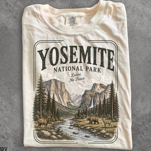 May include: An ivory t-shirt featuring a graphic of Yosemite National Park with the text "YOSEMITE NATIONAL PARK" and "Leave No Trace". The illustration depicts a mountain landscape with a river, pine trees, a deer, and a bear.
