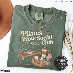 May include: A moss green t-shirt featuring the text "Pilates Flow Social Club" and illustrations of pilates equipment, a coffee cup, and a gym bag. The shirt has a crew neck and short sleeves. A gold necklace is draped across the shirt.