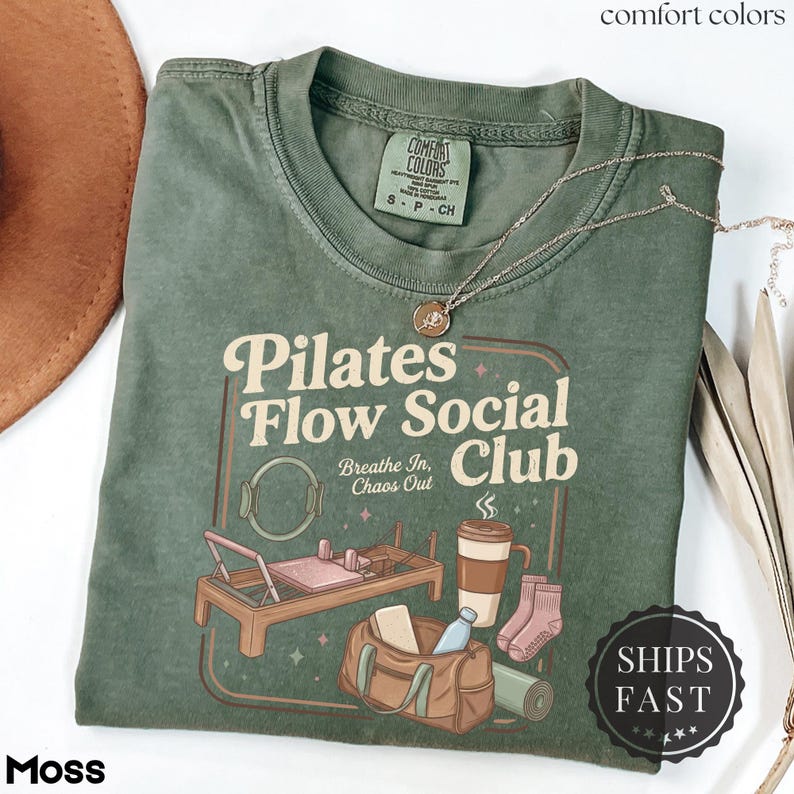 May include: A moss green t-shirt featuring the text "Pilates Flow Social Club" and illustrations of pilates equipment, a coffee cup, and a gym bag. The shirt has a crew neck and short sleeves. A gold necklace is draped across the shirt.