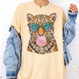 May include: Butter-colored t-shirt featuring a leopard graphic wearing sunglasses and a jeweled necklace, blowing a pink bubble. The leopard's fur is brown and black. The shirt has a relaxed fit.