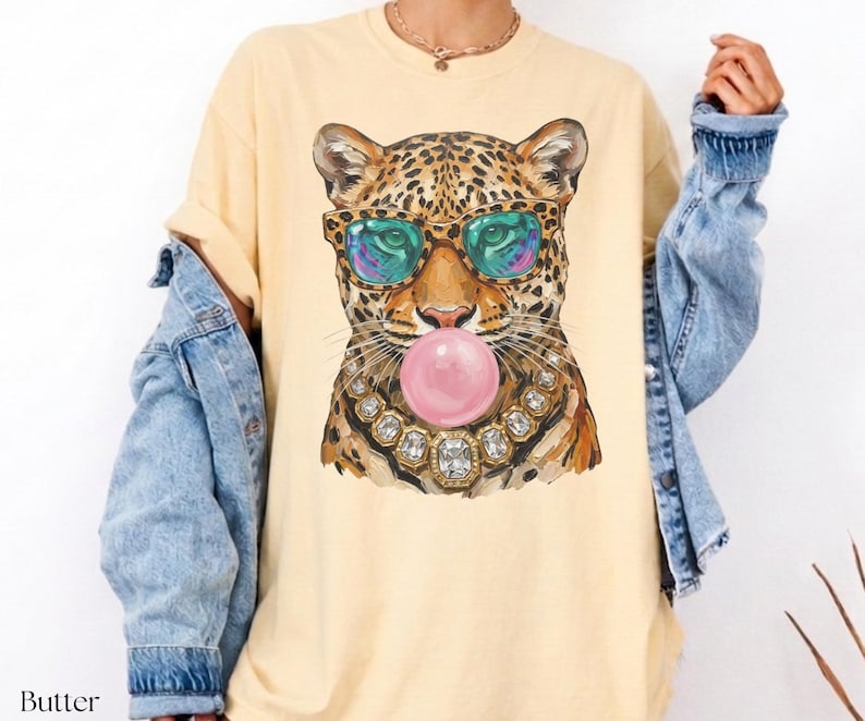May include: Butter-colored t-shirt featuring a leopard graphic wearing sunglasses and a jeweled necklace, blowing a pink bubble. The leopard's fur is brown and black. The shirt has a relaxed fit.