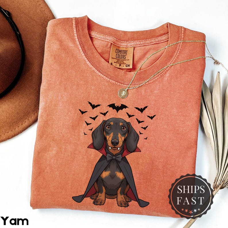 K&ouml;nnte beinhalten: Orangefarbenes Comfort Colors T-Shirt mit einem Dackel als Vampir, mit schwarzem Umhang und Fliege, umgeben von Flederm&auml;usen. Das Shirt hat einen Rundhalsausschnitt und kurze &Auml;rmel. Eine goldene Halskette liegt auf dem Shirt.
