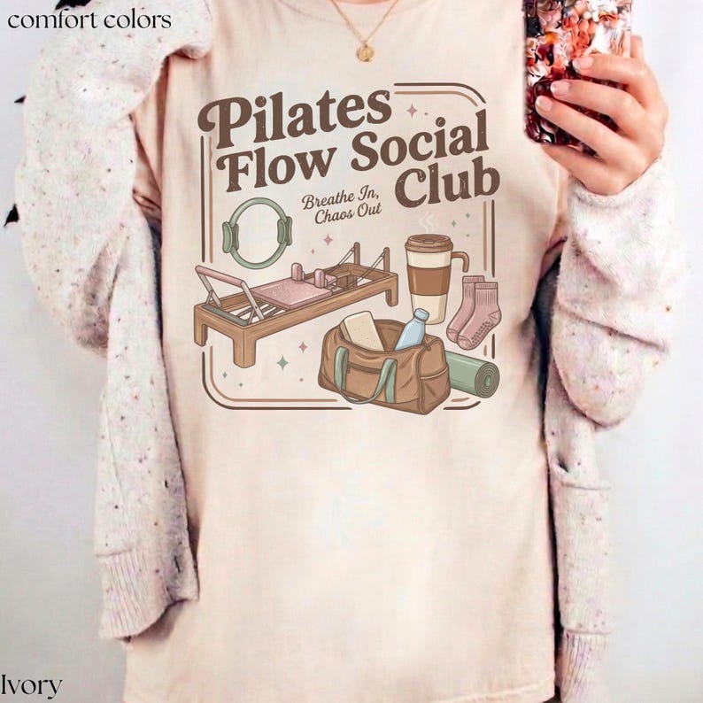 May include: Cream-coloured t-shirt featuring a Pilates Flow Social Club design. The graphic includes a Pilates reformer, a gym bag, a coffee cup, socks, and a yoga mat. The text "Breathe In, Chaos Out" is also included.