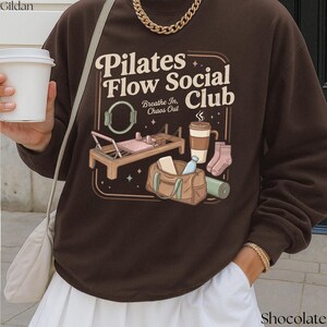 May include: A chocolate brown sweatshirt featuring a graphic for the "Pilates Flow Social Club." The design includes a Pilates reformer, coffee cup, socks, a gym bag, and a yoga mat. The sweatshirt is a Gildan brand.