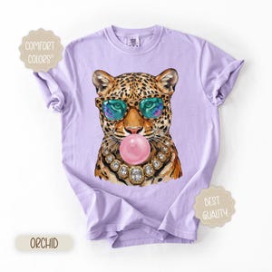 May include: A light purple t-shirt featuring a leopard illustration wearing sunglasses and a jeweled necklace, blowing a pink bubble. The shirt has the words "Comfort Colors" and "Best Quality" on it, and the color is "Orchid".