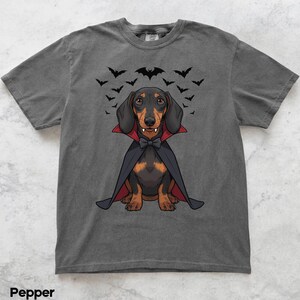K&ouml;nnte beinhalten: Graues T-Shirt mit einem Cartoon-Dackel, der als Vampir verkleidet ist, mit Umhang, Fliege und Fangz&auml;hnen. &Uuml;ber dem Hund fliegt eine Fledermausgruppe. Das T-Shirt ist ein l&auml;ssiger Stil.