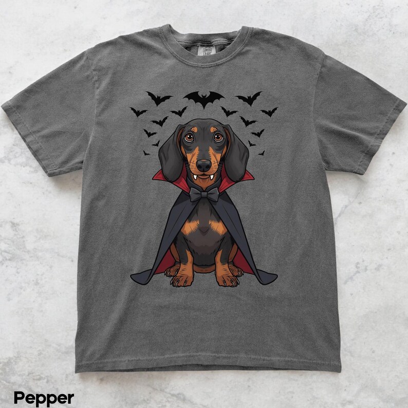 K&ouml;nnte beinhalten: Graues T-Shirt mit einem Cartoon-Dackel, der als Vampir verkleidet ist, mit Umhang, Fliege und Fangz&auml;hnen. &Uuml;ber dem Hund fliegt eine Fledermausgruppe. Das T-Shirt ist ein l&auml;ssiger Stil.