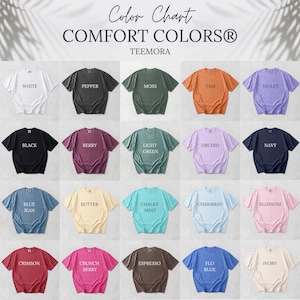 May include: A color chart of Comfort Colors t-shirts in various hues, including white, black, and navy. The chart displays the colors with the names of each color printed on the shirts. The brand name "TEEMORA" is also visible.