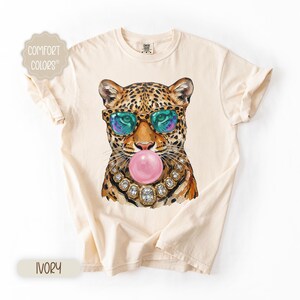 May include: Ivory-colored t-shirt featuring a leopard illustration wearing sunglasses and a jeweled necklace, blowing a pink bubble. The shirt has the words "Comfort Colors" printed on it.