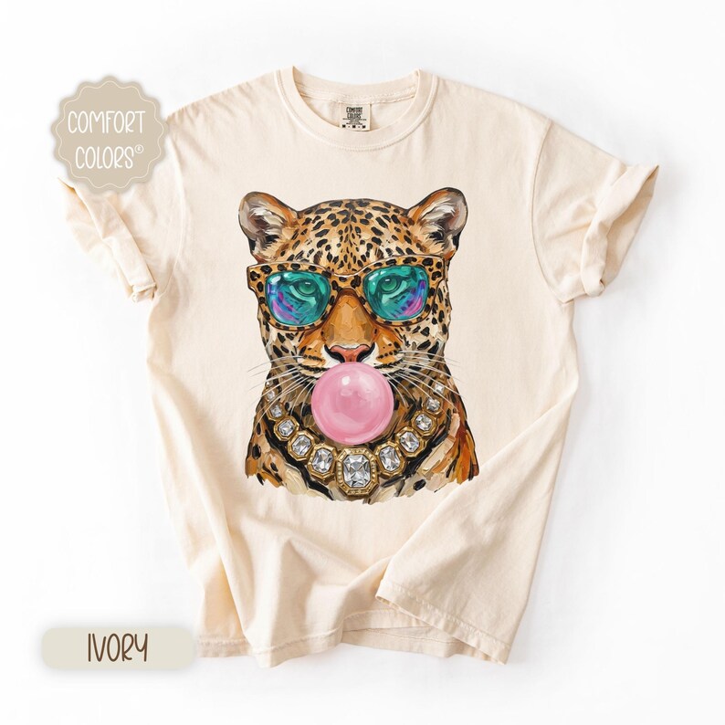 May include: Ivory-colored t-shirt featuring a leopard illustration wearing sunglasses and a jeweled necklace, blowing a pink bubble. The shirt has the words "Comfort Colors" printed on it.