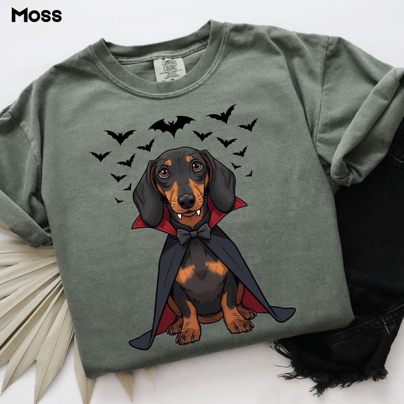 K&ouml;nnte beinhalten: Moosgr&uuml;nes Sweatshirt mit einem Dackel, der als Vampir verkleidet ist, komplett mit schwarzem Umhang, Fliege und Fangz&auml;hnen. Schwarze Flederm&auml;use fliegen &uuml;ber dem Hund. Das Wort "Moss" befindet sich in der oberen linken Ecke.