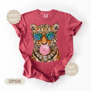 May include: Crimson-colored t-shirt featuring a leopard illustration wearing sunglasses and blowing a bubble. The leopard is adorned with a jeweled necklace. The shirt has the words "Comfort Colors" and "Made for You" printed on it.