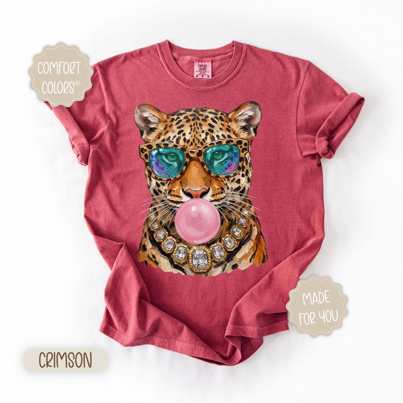 May include: Crimson-colored t-shirt featuring a leopard illustration wearing sunglasses and blowing a bubble. The leopard is adorned with a jeweled necklace. The shirt has the words "Comfort Colors" and "Made for You" printed on it.
