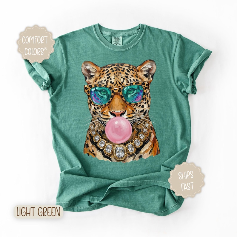 May include: Light green t-shirt featuring a leopard illustration wearing sunglasses and blowing a bubblegum bubble. The leopard is adorned with a jeweled necklace. The shirt has the words "Comfort Colors" and "Light Green" printed on it.