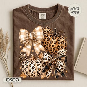 May include: A brown t-shirt featuring a graphic of two pumpkins with leopard print patterns, two large bows with plaid and leopard print designs, and small gold stars. The shirt has a Comfort Colors label at the neck and text reading ALSO IN YOUTH.