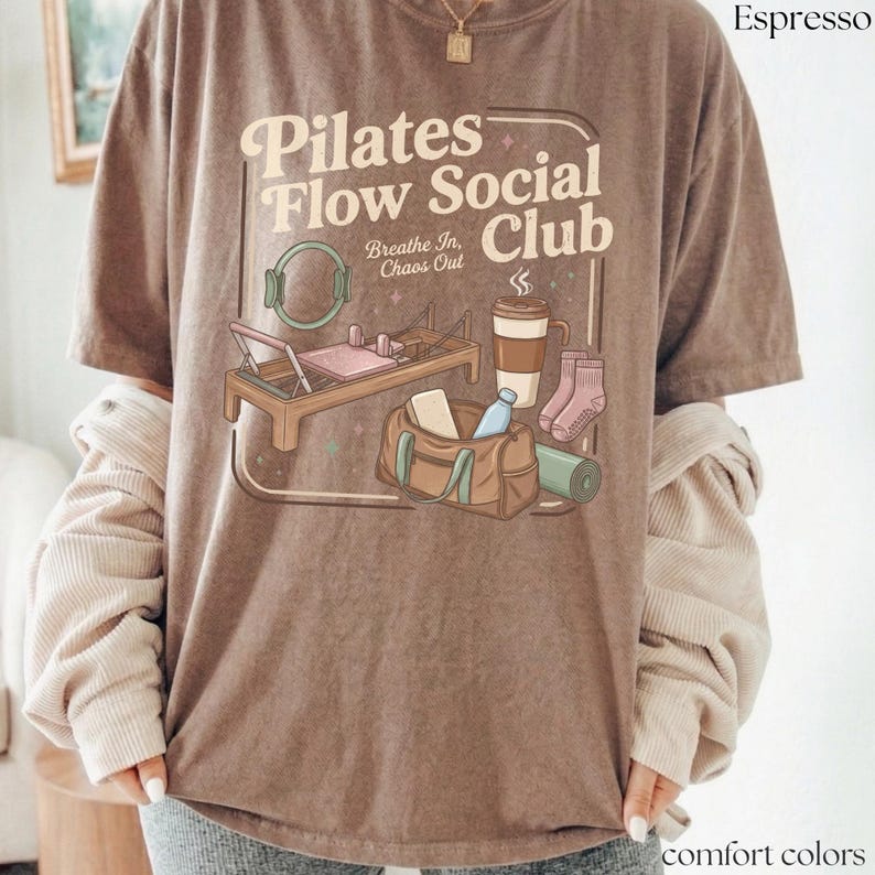 May include: Espresso-coloured t-shirt with the text "Pilates Flow Social Club" and illustrations of pilates equipment, a coffee cup, socks, a gym bag, and a yoga mat. The shirt is a comfort colour.