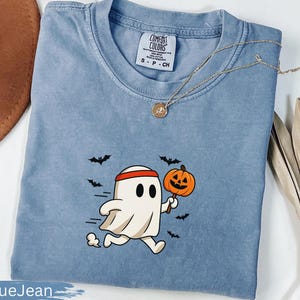 May include: Blue jean colored t-shirt with a cartoon ghost running and holding a jack-o'-lantern. The ghost wears a red headband and is surrounded by black bats. A gold necklace with a pendant is on the shirt.