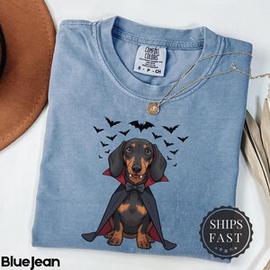 K&ouml;nnte beinhalten: Hellblaues Jeans-T-Shirt mit einem Cartoon-Dackel, der als Vampir verkleidet ist und einen schwarzen Umhang und eine Fliege tr&auml;gt. Das Shirt hat ein Comfort Colors-Etikett und eine Halskette. Schwarze Flederm&auml;use fliegen &uuml;ber dem Hund. Der Text "BlueJean" steht unten.
