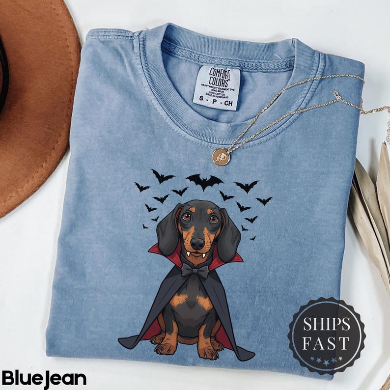 K&ouml;nnte beinhalten: Hellblaues Jeans-T-Shirt mit einem Cartoon-Dackel, der als Vampir verkleidet ist und einen schwarzen Umhang und eine Fliege tr&auml;gt. Das Shirt hat ein Comfort Colors-Etikett und eine Halskette. Schwarze Flederm&auml;use fliegen &uuml;ber dem Hund. Der Text "BlueJean" steht unten.