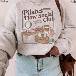 May include: Pale grey sweatshirt featuring a brown graphic with the words "Pilates Flow Social Club". The design includes illustrations of pilates equipment, a coffee cup, socks, a gym bag, and a yoga mat. The text "Breathe In, Chaos Out" is also present.