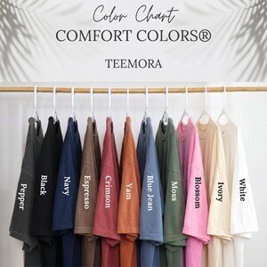 May include: A colour chart showing Comfort Colors t-shirts in a range of shades: Pepper, Black, Navy, Espresso, Crimson, Yam, Blue Jean, Moss, Blossom, Ivory, and White. The shirts are suspended from a wooden rail.