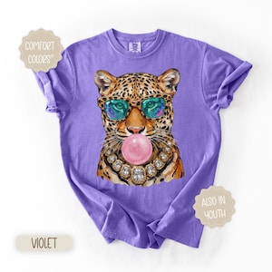 May include: Violet Comfort Colors t-shirt featuring a leopard wearing sunglasses and a jeweled necklace, blowing a pink bubble. The shirt has rolled-up sleeves and the words "Comfort Colors" and "Also in Youth" are printed on the shirt.