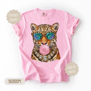 May include: Pink t-shirt featuring a leopard wearing sunglasses and a jeweled necklace, blowing a bubblegum bubble. The shirt has the words "Comfort Colors", "Blossom", and "Also in Youth" printed on it.