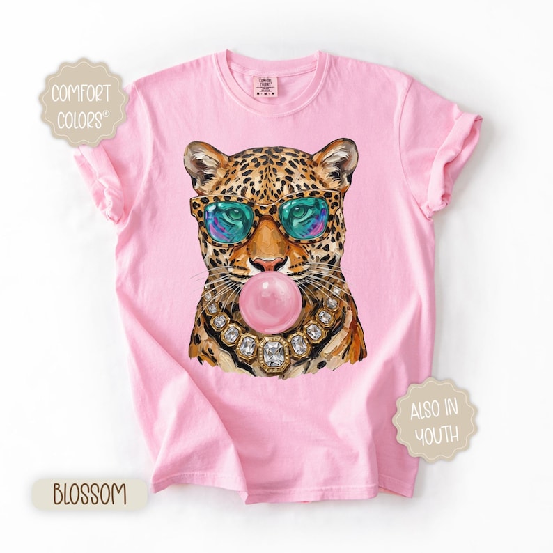 May include: Pink t-shirt featuring a leopard wearing sunglasses and a jeweled necklace, blowing a bubblegum bubble. The shirt has the words "Comfort Colors", "Blossom", and "Also in Youth" printed on it.