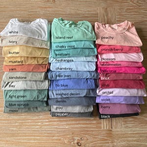 K&ouml;nnte beinhalten: Gestapelte T-Shirts in verschiedenen Farben, darunter Wei&szlig;, Elfenbein, Butter, Senf und Sandstein. Weitere Farben sind Island Reef, Kreideminze, Meerschaum und Hortensie. Die Shirts sind ordentlich gefaltet und farblich beschriftet.