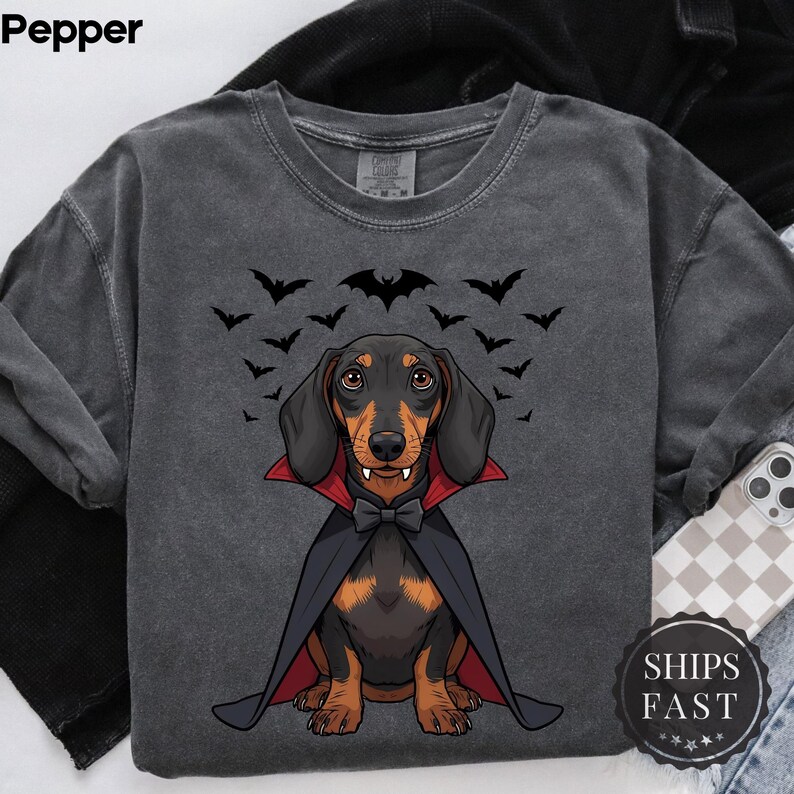 K&ouml;nnte beinhalten: Dunkelgraues Sweatshirt mit einem Dackel als Vampir, mit schwarzem Umhang und Fliege, umgeben von Flederm&auml;usen. Das Wort "Pepper" steht oben links. Der Text "SHIPS FAST" steht unten rechts.