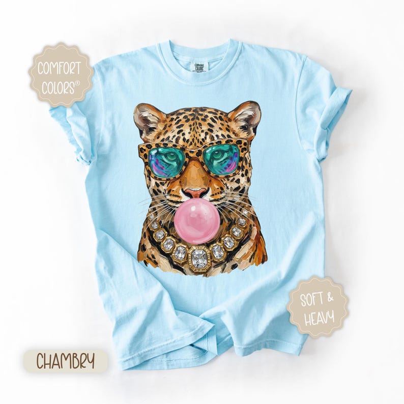 May include: Light blue t-shirt featuring a leopard illustration wearing sunglasses and blowing a bubblegum bubble. The leopard is adorned with a jeweled necklace. The shirt has the words "Comfort Colors", "Chambry", and "Soft & Heavy" printed on it.