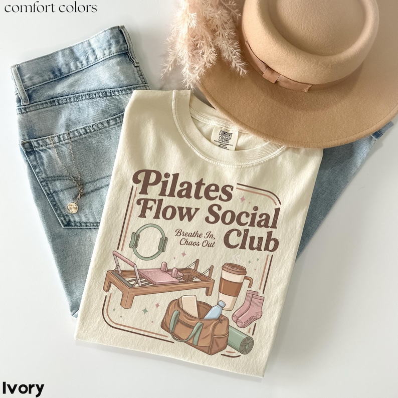 May include: Cream-coloured t-shirt featuring the text "Pilates Flow Social Club" and illustrations of pilates equipment, a coffee cup, and a gym bag. The shirt is styled with light-wash jeans, a necklace, and a beige hat.