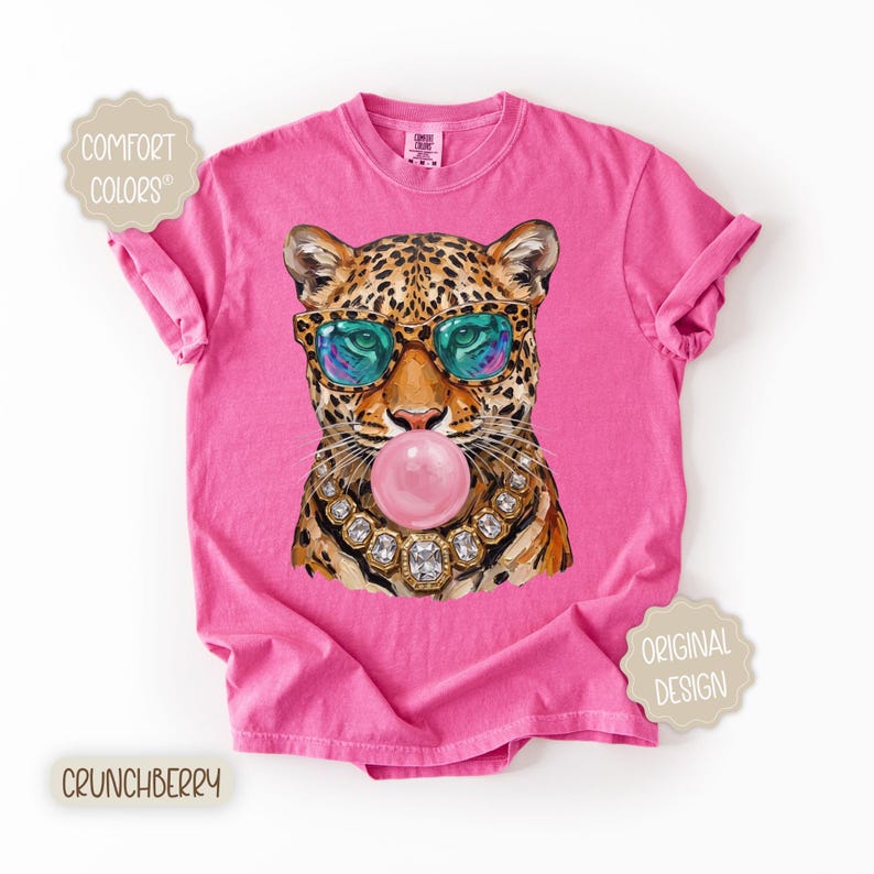 May include: Pink t-shirt featuring a leopard wearing sunglasses and a jeweled necklace, blowing a bubblegum bubble. The shirt has the words "Comfort Colors", "Original Design", and "Crunchberry" printed on it.