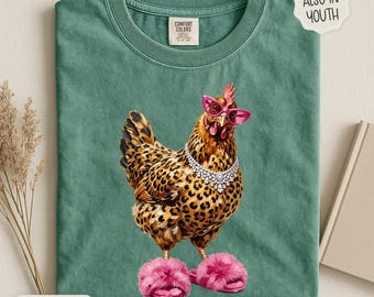 May include: A sage green t-shirt featuring a graphic of a chicken with leopard print feathers, wearing bright pink cat-eye glasses and a silver rhinestone necklace. The chicken is also wearing fluffy pink slippers. The shirt has a neck label.