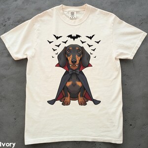 K&ouml;nnte beinhalten: Elfenbeinfarbenes T-Shirt mit einem Cartoon-Dackel, der als Vampir verkleidet ist, komplett mit Umhang, Fliege und Fangz&auml;hnen. Schwarze Flederm&auml;use fliegen &uuml;ber dem Hund. Das Shirt hat einen Rundhalsausschnitt und kurze &Auml;rmel.