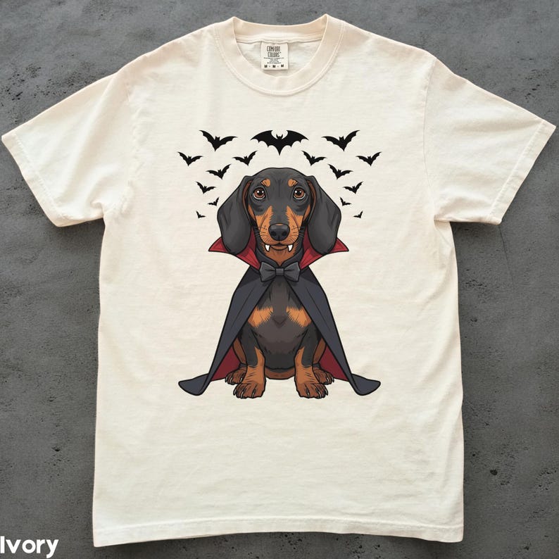 K&ouml;nnte beinhalten: Elfenbeinfarbenes T-Shirt mit einem Cartoon-Dackel, der als Vampir verkleidet ist, komplett mit Umhang, Fliege und Fangz&auml;hnen. Schwarze Flederm&auml;use fliegen &uuml;ber dem Hund. Das Shirt hat einen Rundhalsausschnitt und kurze &Auml;rmel.