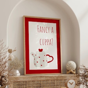 Könnte beinhalten: Ein gerahmter Druck mit rotem Rand zeigt einen weißen Hintergrund mit einer Teekannenillustration. Die Teekanne ist mit roten Herzen verziert und hat einen roten Griff. Der Text "FANCY A CUPPA?" steht in Rot über der Teekanne.