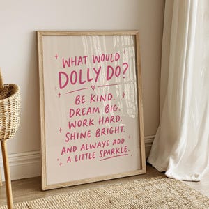 May include: A rectangular framed wall art piece with a light beige background and pink text that reads: WHAT WOULD DOLLY DO? BE KIND. DREAM BIG. WORK HARD. SHINE BRIGHT. AND ALWAYS ADD A LITTLE SPARKLE. The frame is made of light-coloured wood.